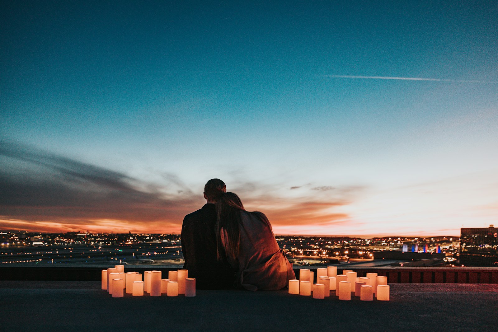 Couple sitting together behind rows of glowing candles, looking out over a city at dusk
