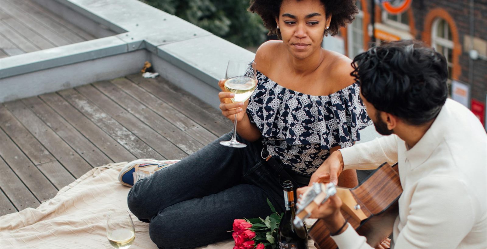 Couple sharing wine on a rooftop picnic date with a guitar and flowers